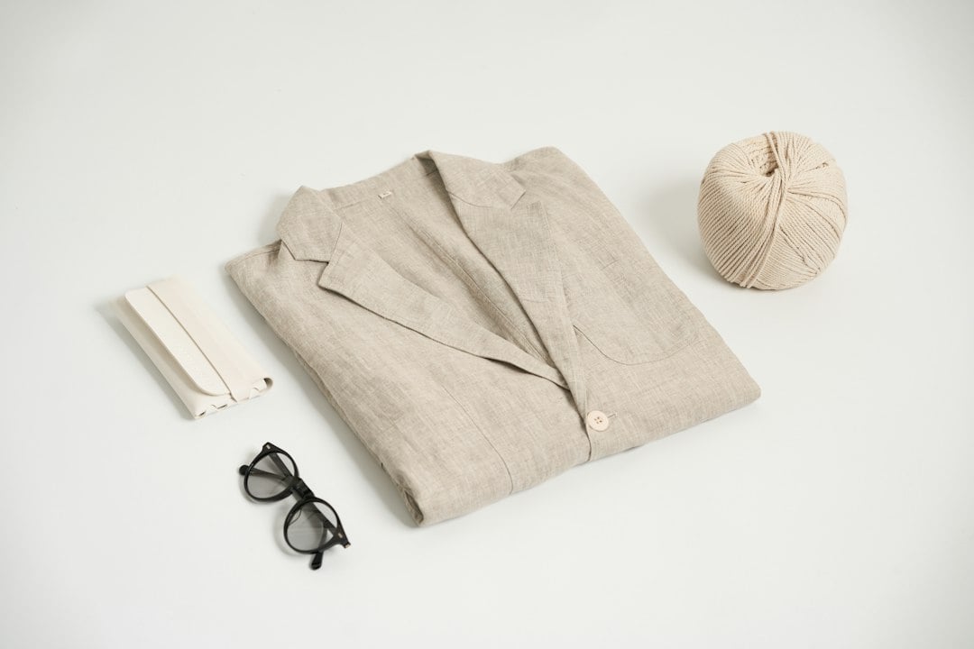 A beige linen shirt laid flat on a white surface beside folded fabric, black-rimmed glasses and a ball of cream twine.