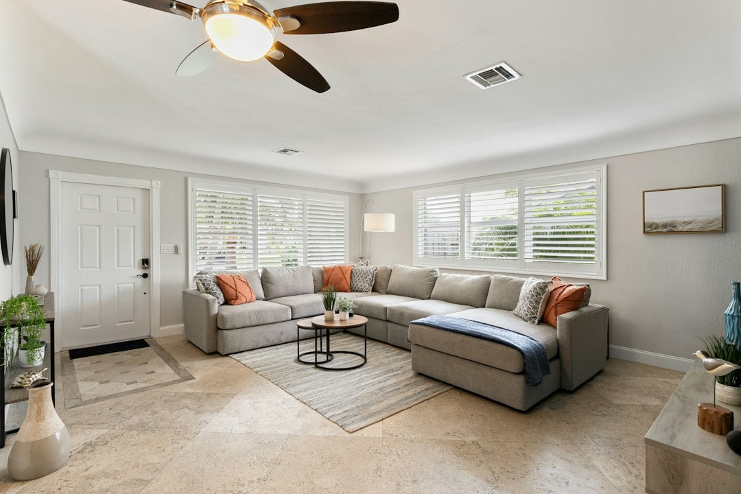 Sunlit vacation rental living room with a beige sectional sofa, ceiling fan and shuttered windows