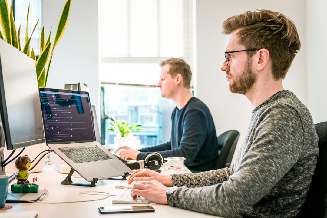 Two men work at office desks with laptops on stands and external monitors.