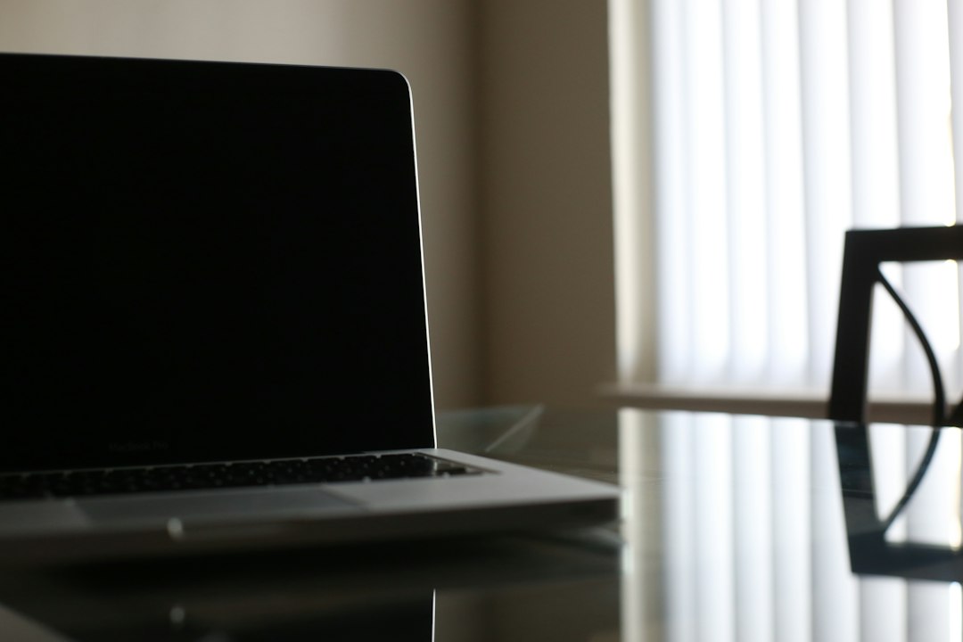 An open silver laptop with a dark screen sits on a glass table next to a bright window.