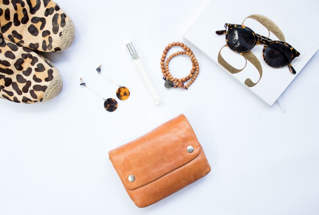 White flat lay of a leopard-print shoe, tan leather wallet, sunglasses, bracelets and a slim clear fragrance vial
