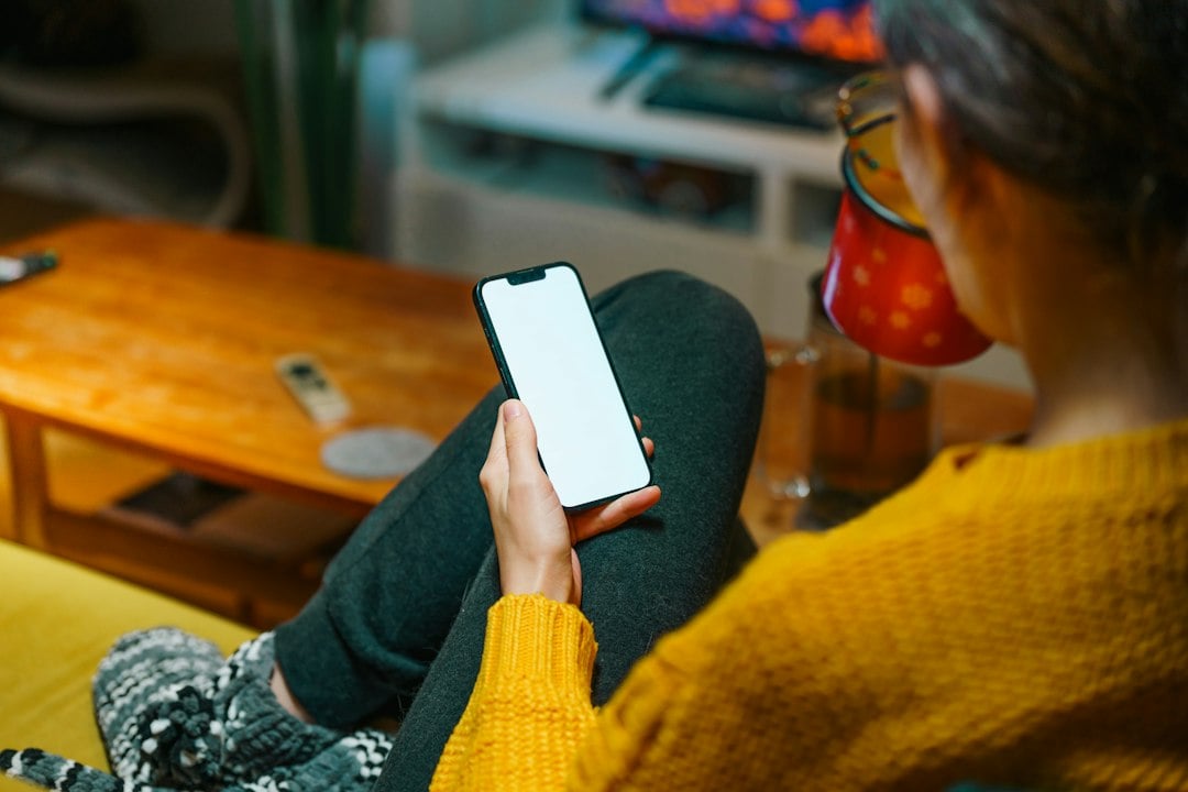 Over-the-shoulder view of a woman in a yellow jumper using a phone with a blank screen on her sofa