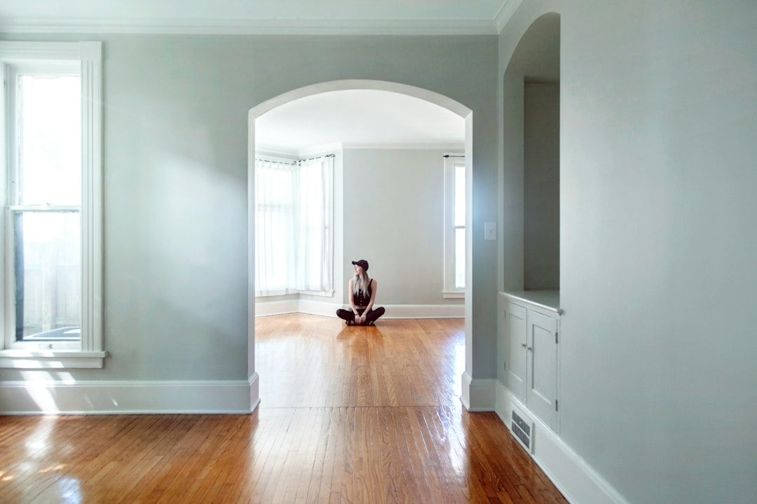 A person sits alone on the hardwood floor of an empty home framed by an arched doorway