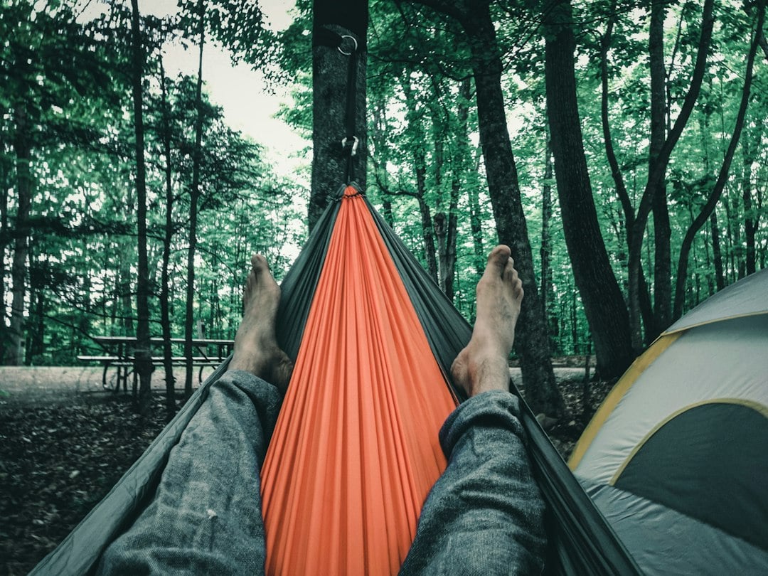 Point-of-view shot of bare feet crossed in an orange hammock at a wooded campsite with a tent alongside