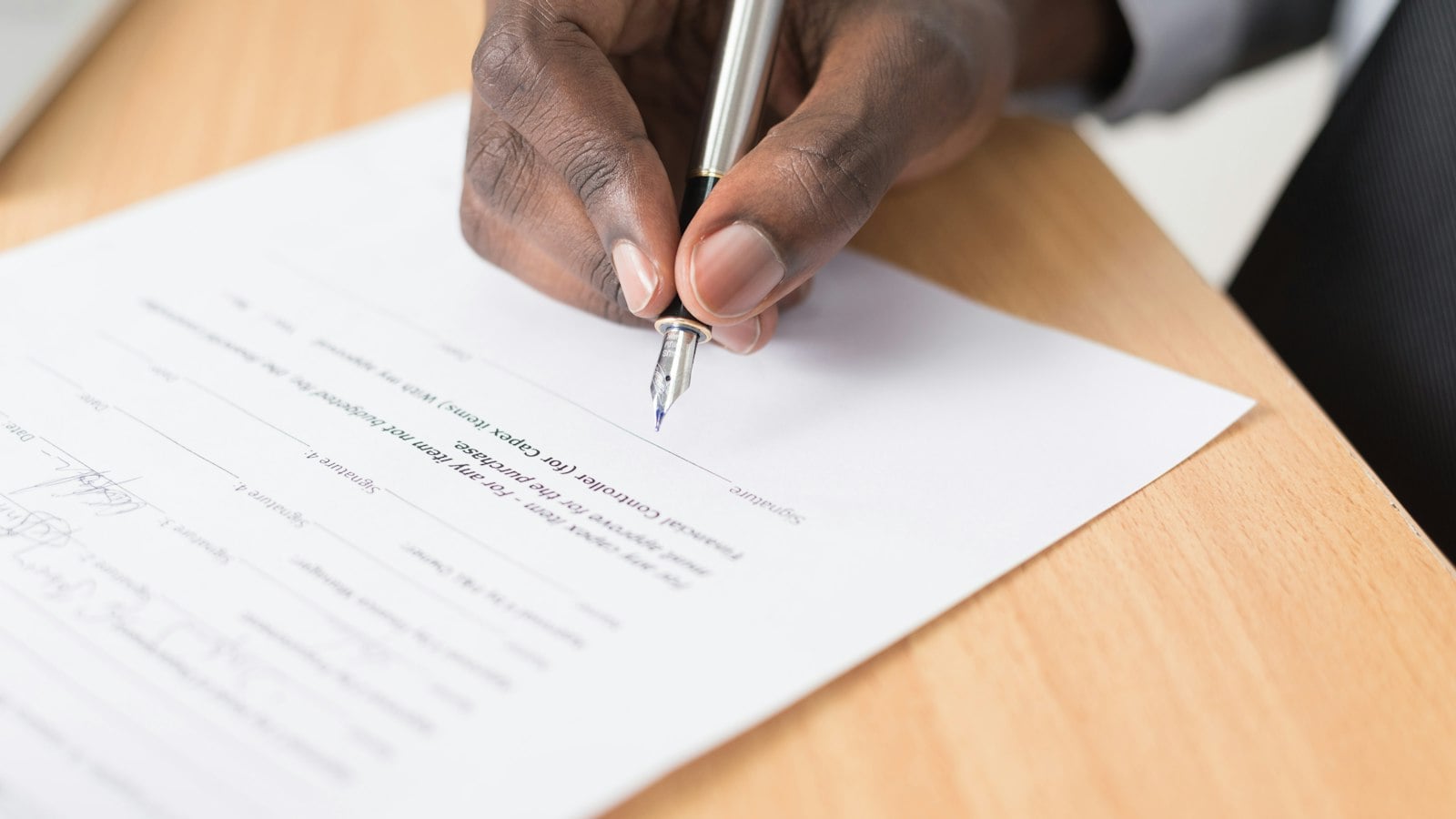 A hand signing a printed document with a pen on a wooden desk (photo: Cytonn Photography / Unsplash)