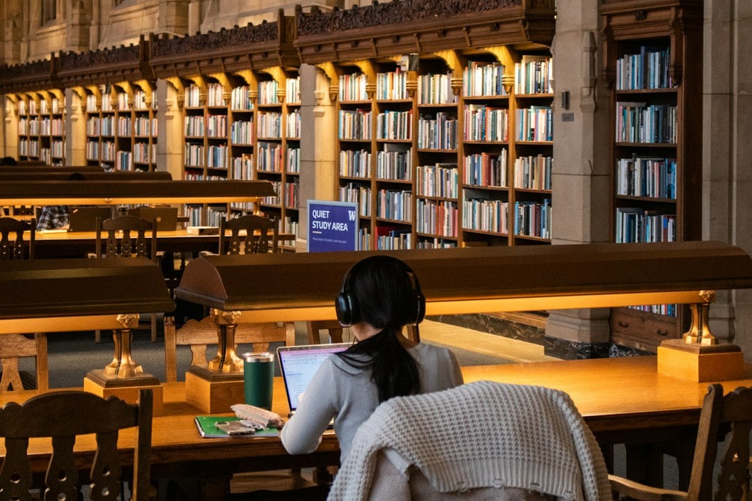 A student wearing headphones works at a laptop at a long table in a grand library reading room.