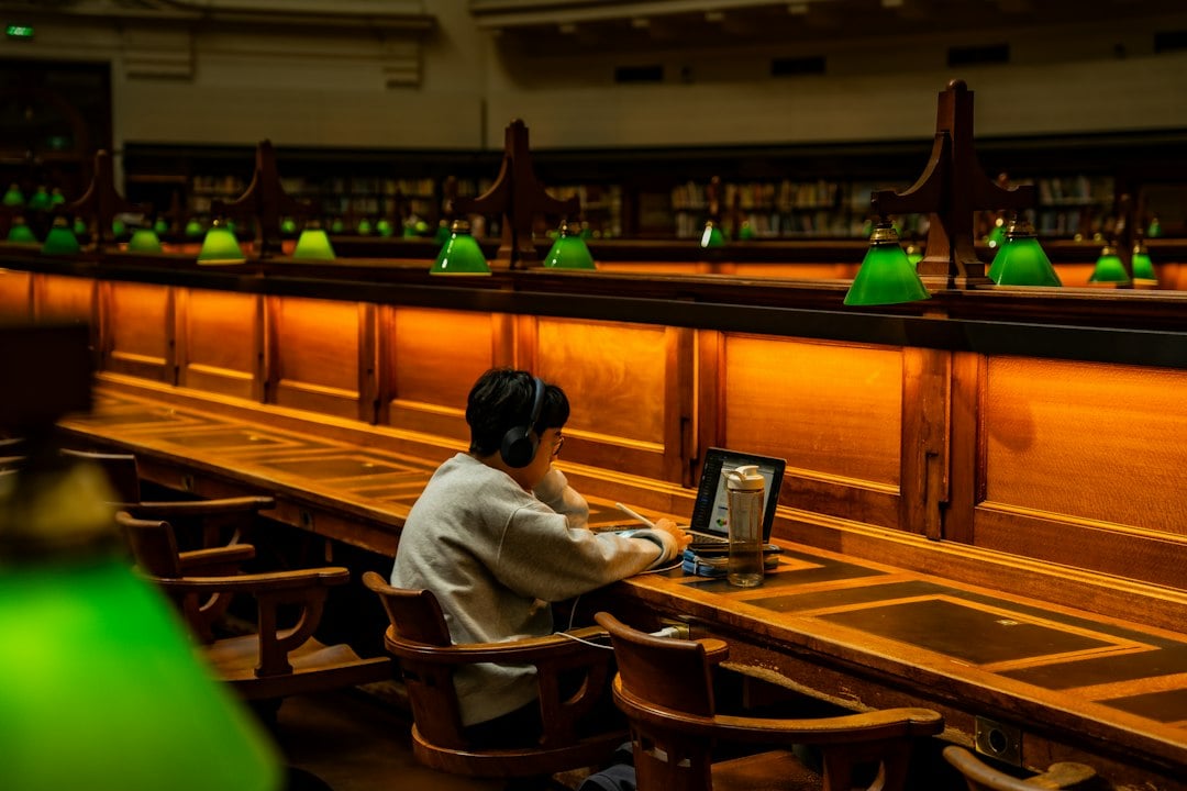 A lone student works at a laptop in a dark reading room lined with green desk lamps.