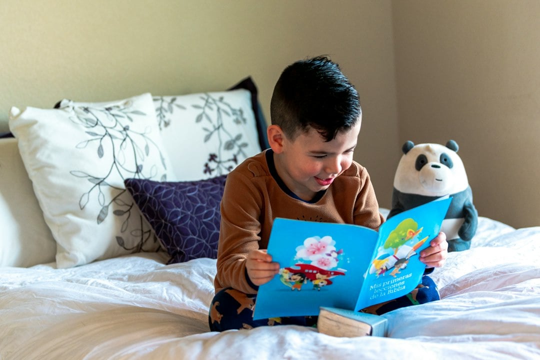 A smiling boy reads a picture book on a bed with a panda plush toy next to him