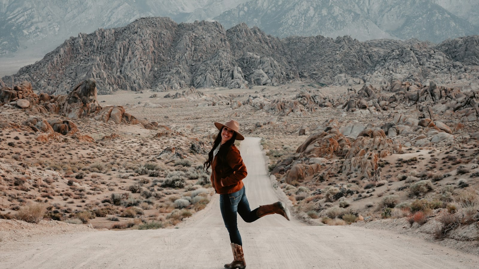 A woman in a wide-brimmed hat walking along a dirt road through desert scrub and rocky hills (photo: Royce Fonseca / Unsplash)