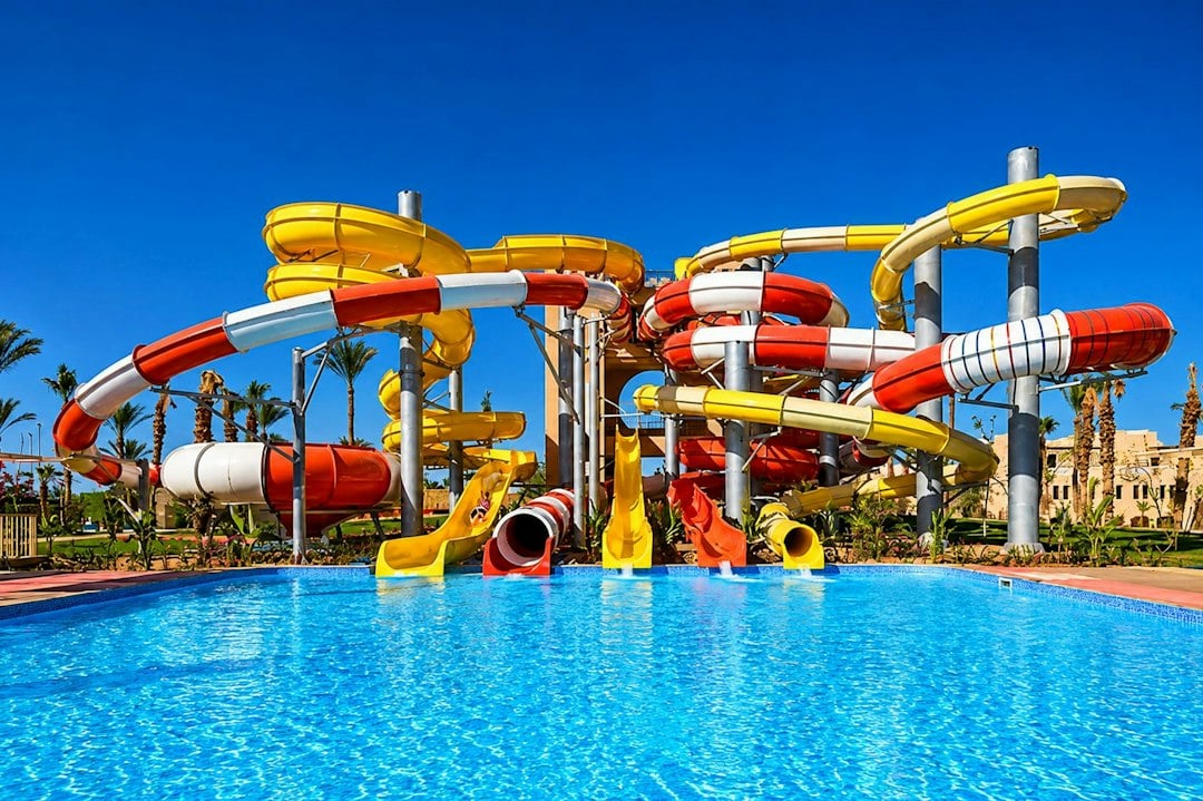 Tall red and yellow water slides tower over a bright blue resort pool on a sunny day