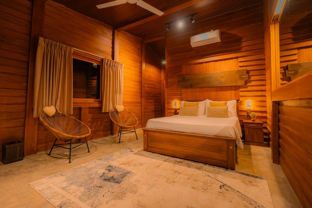 A wood-paneled resort bedroom with a king bed, two rattan chairs and a patterned rug